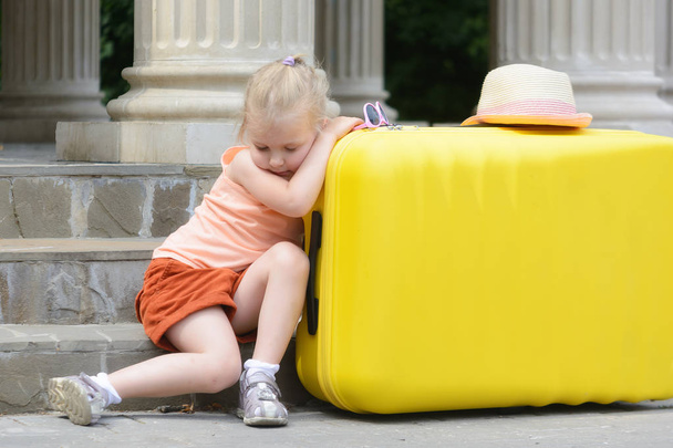 Маленька дівчинка заснула, нахиливши лікті на велику жовту валізу. Портрет красивої дівчини. Фон розмитий
. - Фото, зображення