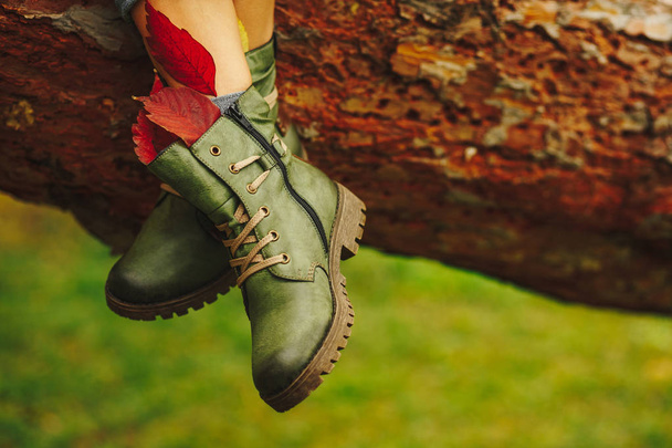 Botas de cuero verde en las piernas de las mujeres decoradas con hojas de otoño de follaje sobre fondo de árbol marrón. Diseño, concepto de calzado, publicidad
. - Foto, Imagen