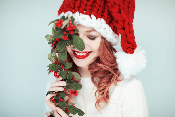 Santas little helper. Beautiful happy young woman with a santa claus hat, perfect make up, red lipstick, holding branch with red berries - Photo, image