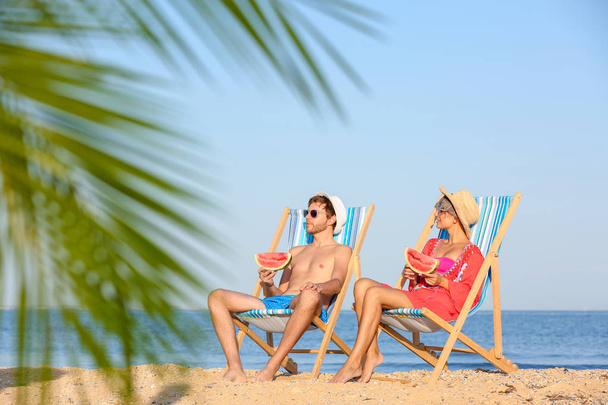Молодая пара с ломтиками арбуза в пляжных креслах на берегу моря
 - Фото, изображение