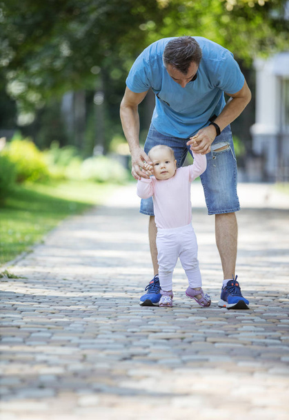Отец поддерживает маленькую дочь и помогает ей сделать первые шаги. Папа и дочь на прогулке в летнем парке
. - Фото, изображение