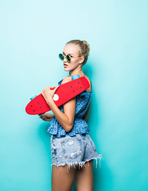 Αίσθηση ελεύθερη και ευτυχής. Ελκυστική νεαρή γυναίκα, χαμογελαστά και μεταφέρουν skateboard ενώ στέκεται ενάντια σε μπλε φόντο - Φωτογραφία, εικόνα