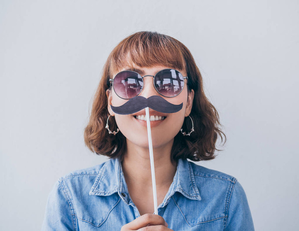 Beautiful asian woman short hair wearing blue jeans shirt and sunglasses smiling with fake mustache isolated on white background. - Photo, Image