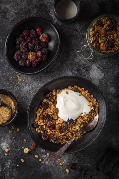 homemade granola (hazelnuts, raisin, dried cranberry, pumpkin, sesame and sunflower seeds) with yogurt in black bowl. Top view. Dark background - Φωτογραφία, εικόνα