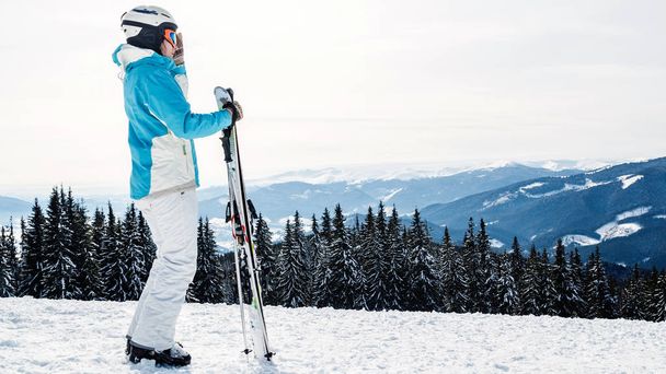 青いスーツ、ヘルメット、眼鏡の女性はスキー山の上に立っています。. - 写真・画像