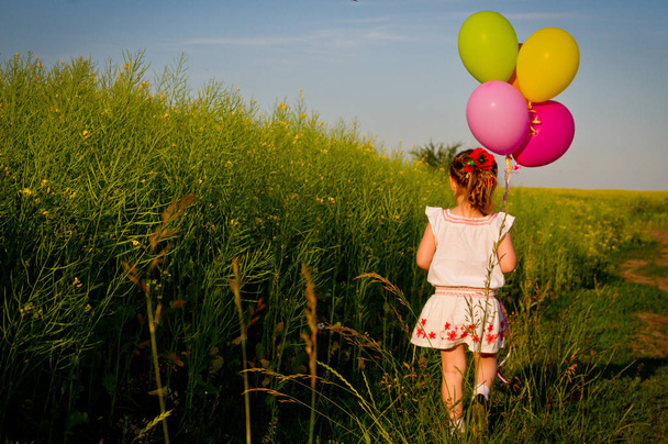 beetje Aziatisch meisje in een jurk loopt door groene tarweveld met ballonnen in de hand, slow-motion - Foto, afbeelding