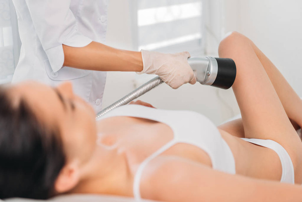 vista parcial del cosmetólogo haciendo masaje eléctrico de pierna a mujer en ropa interior blanca en el salón de spa
 - Foto, imagen