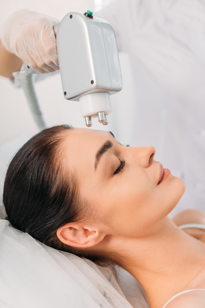 visión parcial de la mujer recibiendo tratamiento facial de microcorriente hecho por el cosmetólogo en el salón de spa
 - Foto, Imagen