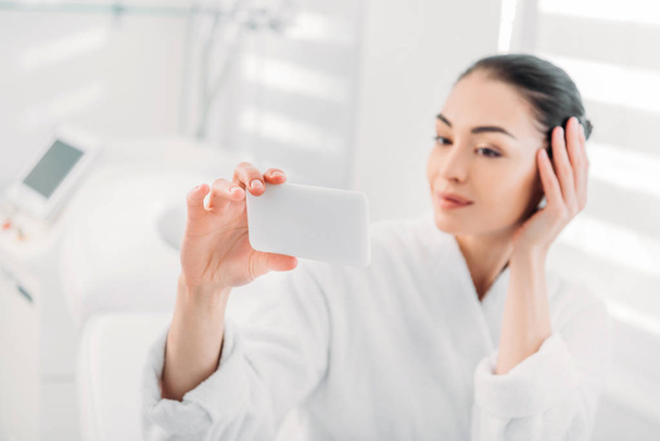 woman in white bathrobe taking selfie on smartphone in spa salon - Foto, Bild