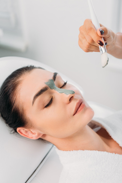 částečný pohled kosmetička použití jílová maska na ženské tváře ve spa salonu - Fotografie, Obrázek