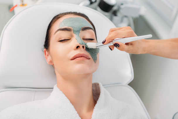 visão parcial de cosmetologist aplicando máscara de barro no rosto feminino no salão de spa
 - Foto, Imagem