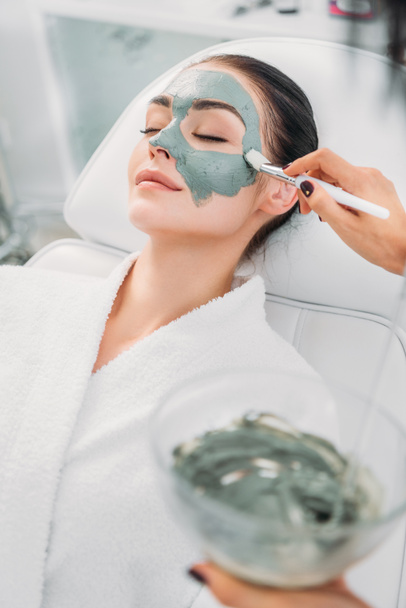 vista parziale di cosmetologo che applica maschera di argilla su faccia femminile in salone di centro termale
 - Foto, immagini
