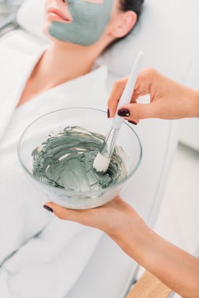 selective focus of cosmetologist with clay mask in bowl and brush in spa salon - Foto, Imagen