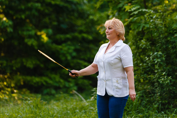 senior woman holding badminton racket standing in green park - Foto, Imagem