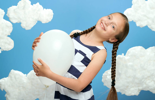 girl dressed in striped dress posing on a blue background with cotton clouds, white air balloon, the concept of summer and happiness - Photo, Image