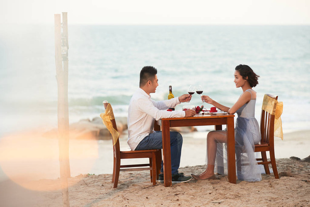 Asian couple holding hands and toasting with red wine while celebrating, picturesque seaside of tropical island - Photo, Image