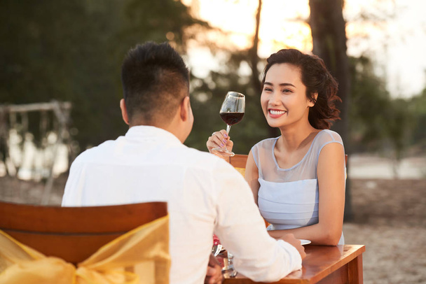 Ελκυστική νεαρή γυναίκα με γοητευτικό χαμόγελο αναπνέοντας κόκκινο κρασί και ακούγοντας το αγαπώντας φίλο με ενδιαφέρον ξοδεύοντας το βράδυ στο εστιατόριο beach - Φωτογραφία, εικόνα