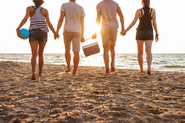 Cropped back view photo of group of friends loving couples walking outdoors on the beach having fun holding boxes with food and drinks. - Foto, Bild
