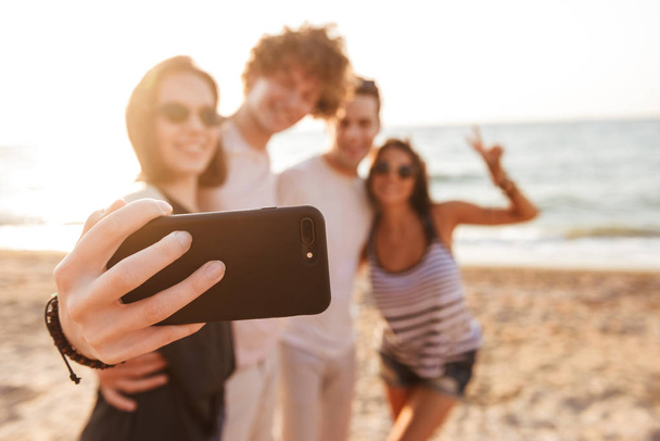 Photo of group of happy friends loving couples outdoors on the beach take a selfie by mobile phone with peace gesture. - Foto, afbeelding