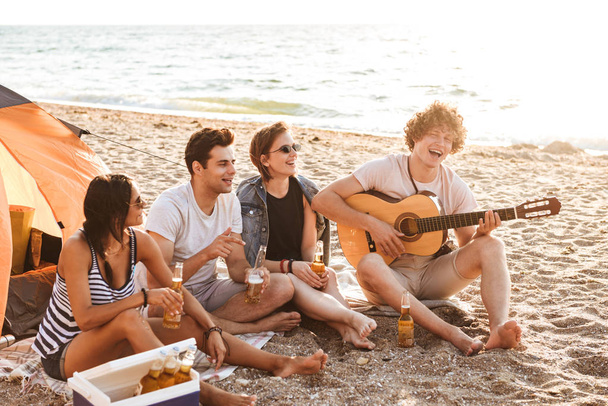 Group of excited young friends having fun time together at the beach, drinking beer, playing guitar while camping - Φωτογραφία, εικόνα