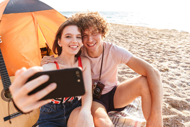 Happy young couple sitting together at the beach, camping, taking selfie - Φωτογραφία, εικόνα