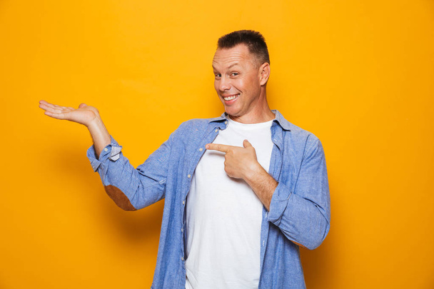 Portrait of a cheerful middle aged man pointing finger, presenting copy space isolated over yellow background - Foto, afbeelding