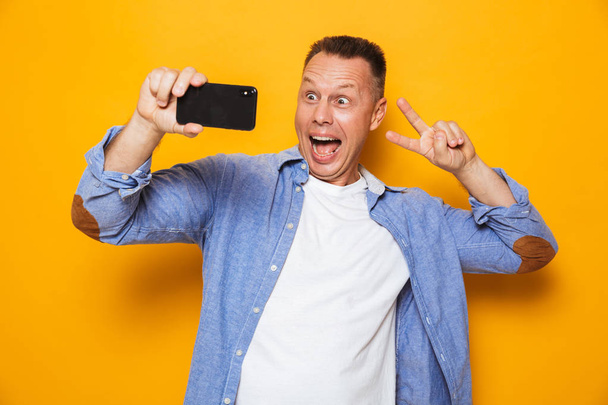 Portrait of a happy middle aged man taking a selfie, showing peace isolated over yellow background - Foto, afbeelding