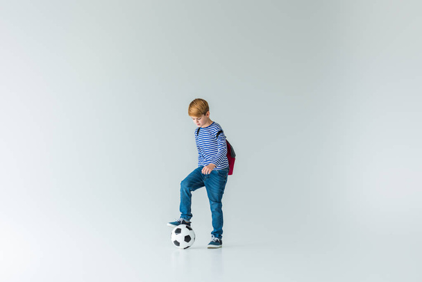 Rozkošný školák s batohem dát nohu na fotbalové míče na bílém - Fotografie, Obrázek
