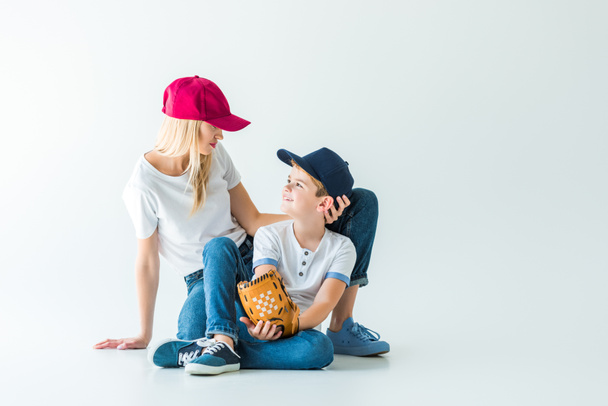 mother and son in caps sitting on floor with baseball glove on white and looking at each other - Foto, Imagem