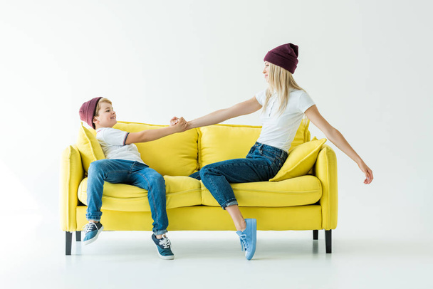 side view of mother and son in burgundy hats holding hands on yellow sofa on white - Photo, Image