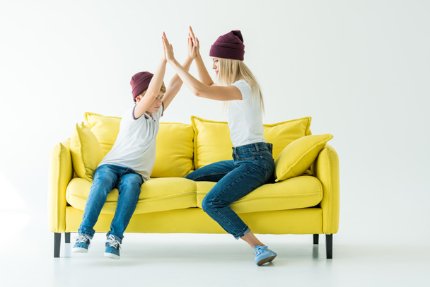 mother and son in burgundy hats giving high fives on yellow sofa on white - Fotoğraf, Görsel