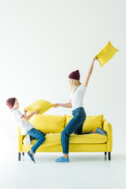 mother and son in burgundy hats having fun and fighting with cushions on yellow sofa on white - Photo, Image