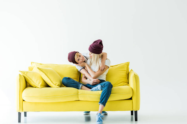 mother and son in burgundy hats hugging on yellow sofa on white - Foto, Imagem