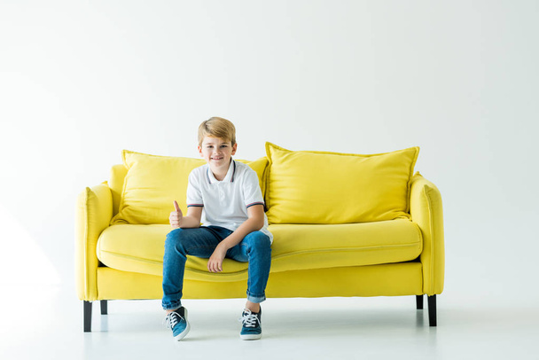 smiling adorable boy sitting on yellow sofa and showing thumb up on white - Fotó, kép