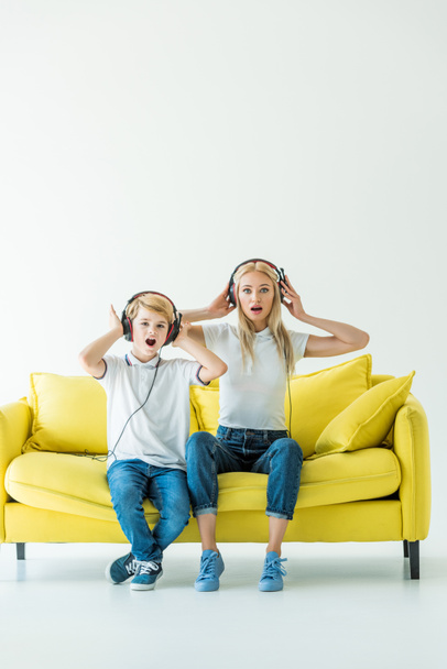 shocked mother and son listening music with headphones and touching heads on yellow sofa on white - Foto, immagini