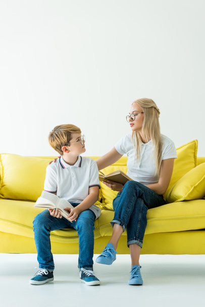 mother and son in glasses holding books and looking at each other on yellow sofa - Фото, изображение
