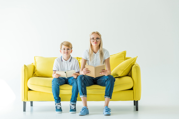 smiling mother and son in glasses holding books and looking at camera on yellow sofa - 写真・画像