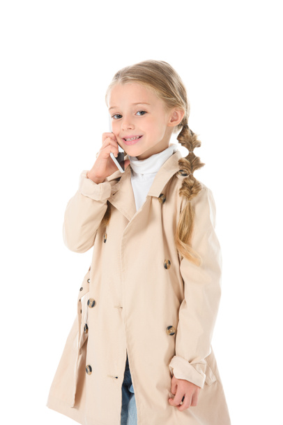 чарівна дитина в осінньому бежевому пальто розмовляє на смартфоні, ізольована на білому
 - Фото, зображення
