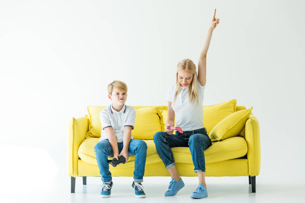 mother showing one hand up after winning video game, son sitting upset on yellow sofa on white - 写真・画像
