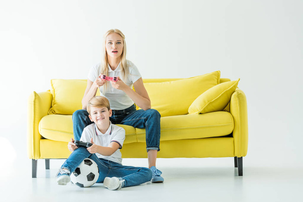 excited mother and son playing video game on yellow sofa on white, football ball on floor - Fotografie, Obrázek
