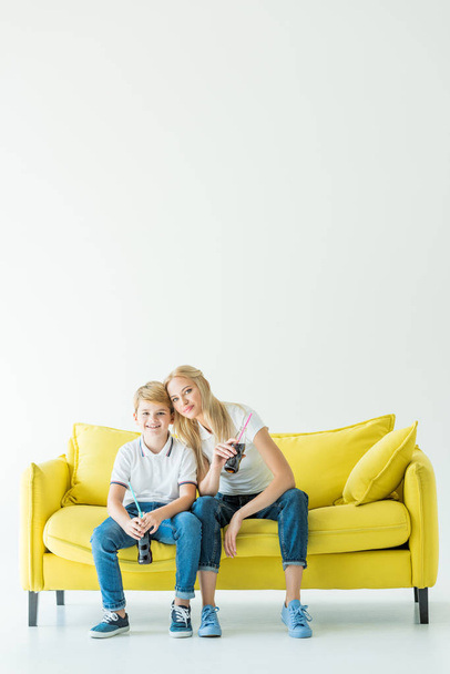 smiling mother and son holding bottles of soda and sitting on yellow sofa - Foto, afbeelding