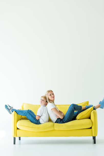 μητέρα και γιος κάθεται με σταυρωμένα τα χέρια στον κίτρινο καναπέ και να βλέπουν τα φωτογραφικών μηχανών σε λευκό - Φωτογραφία, εικόνα