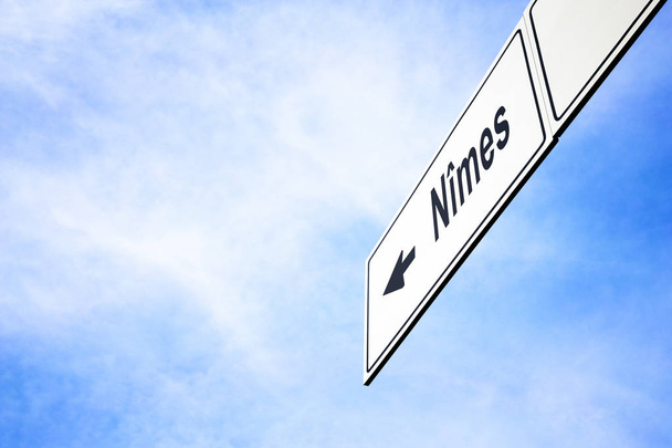 Λευκή πινακίδα με ένα βέλος που δείχνει αριστερά προς Nimes, Γαλλία, ενάντια σε ένα θολό μπλε του ουρανού σε μια έννοια του ταξιδιού, πλοήγησης και κατεύθυνσης. Διαδρομή που περιλαμβάνονται για την πινακίδα - Φωτογραφία, εικόνα