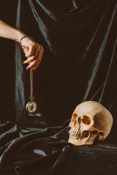 cropped view of woman holding vintage clock on the chain near skull on black cloth - Fotó, kép
