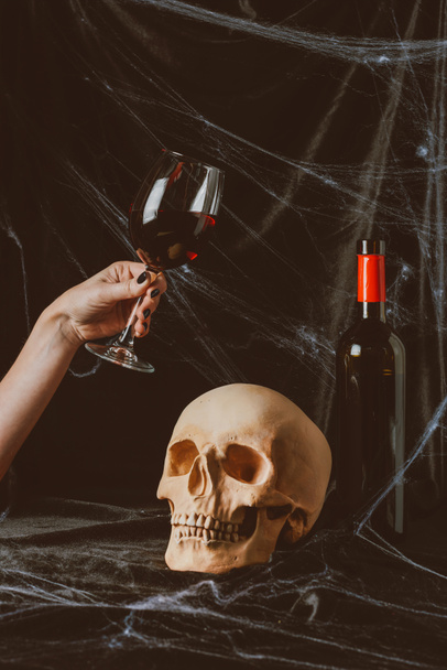 cropped view of woman holding glass of red wine near skull on black cloth with spider web - 写真・画像