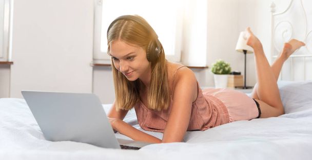 Woman with headphones listening to the music and browsing internet on bed in bedroom. - Foto, afbeelding