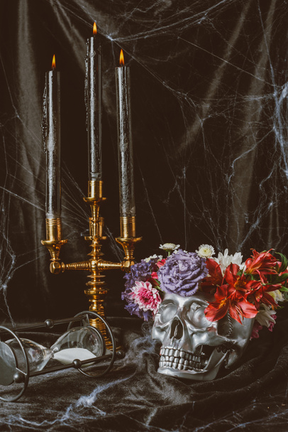 hourglass, silver skull with flowers and candelabrum with candles on black cloth with spider web, decorations for halloween - Photo, Image