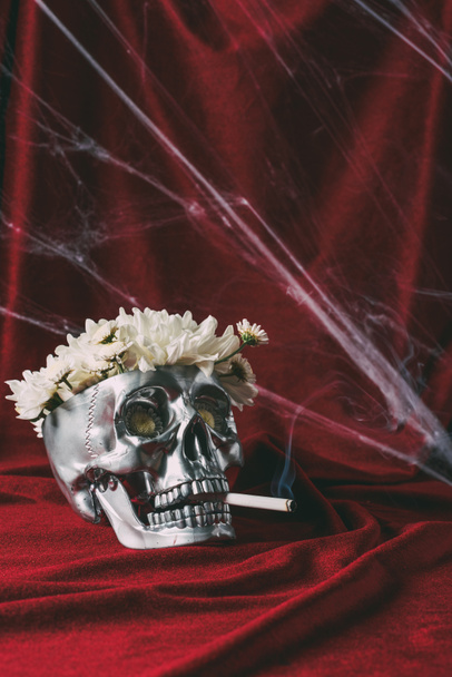 crâne argenté avec des fleurs fumant cigarette sur tissu rouge avec toile d'araignée
  - Photo, image