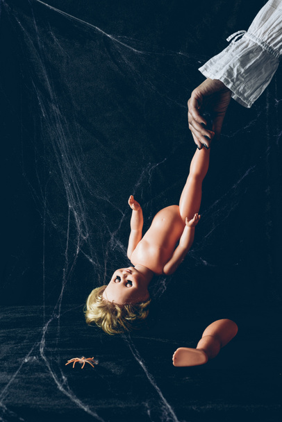vista parcial de mujer asustadiza sosteniendo muñeca espeluznante en la oscuridad con tela de araña
 - Foto, Imagen