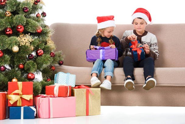 brother and sister in santa hat holding presents and sitting on sofa near christmas tree, isolated on white - Photo, image